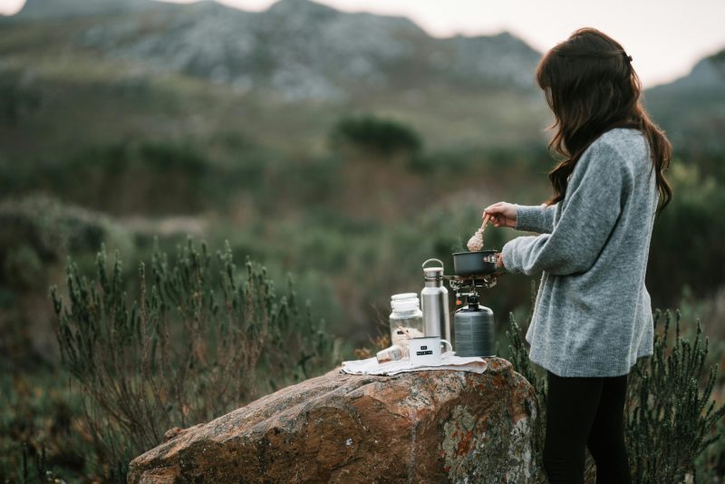 Une femme prépare un petit-déjeuner chaud sur un réchaud de camping portable au milieu d'un cadre naturel pittoresque.