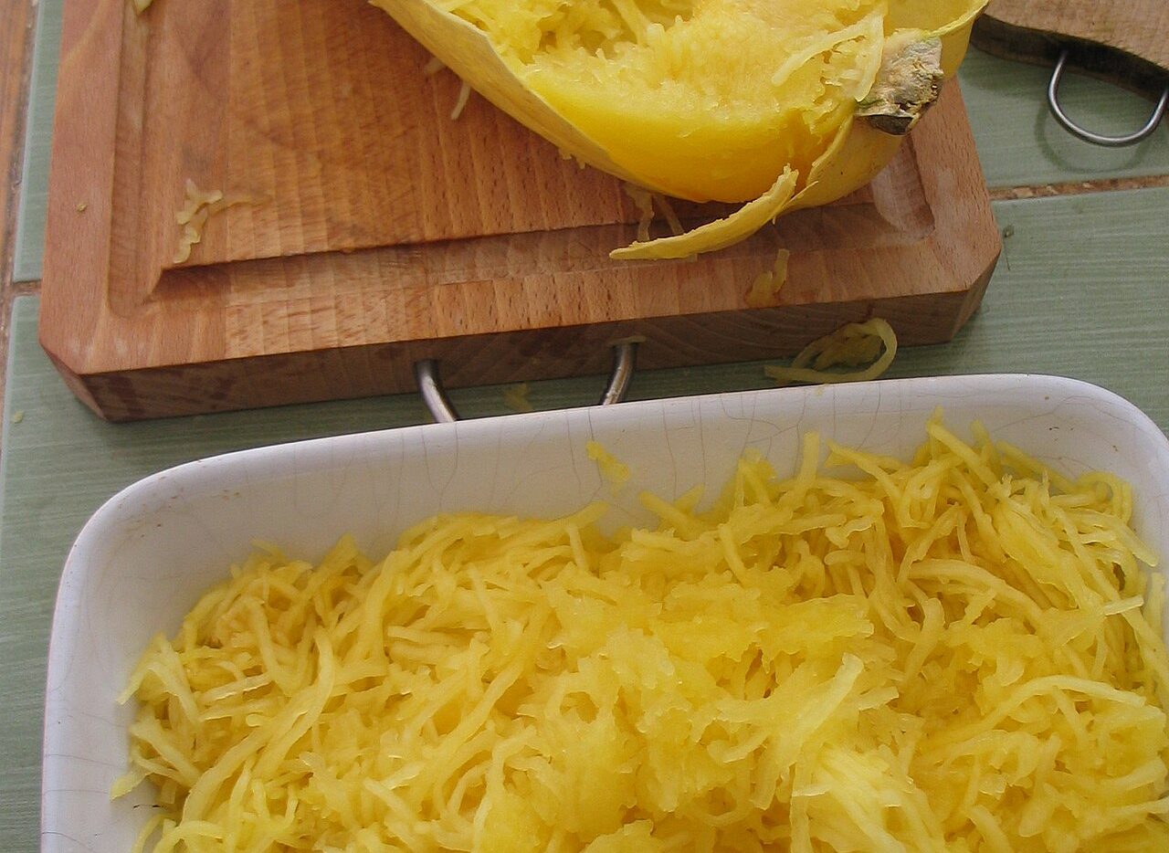 Moitié de courge spaghetti cuite sur une planche, filaments jaunes détachés à la fourchette et plat de filaments