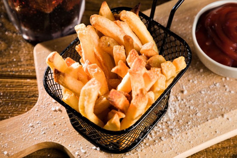 Frites dorées dans un panier métallique noir posé sur une planche, avec une sauce rouge